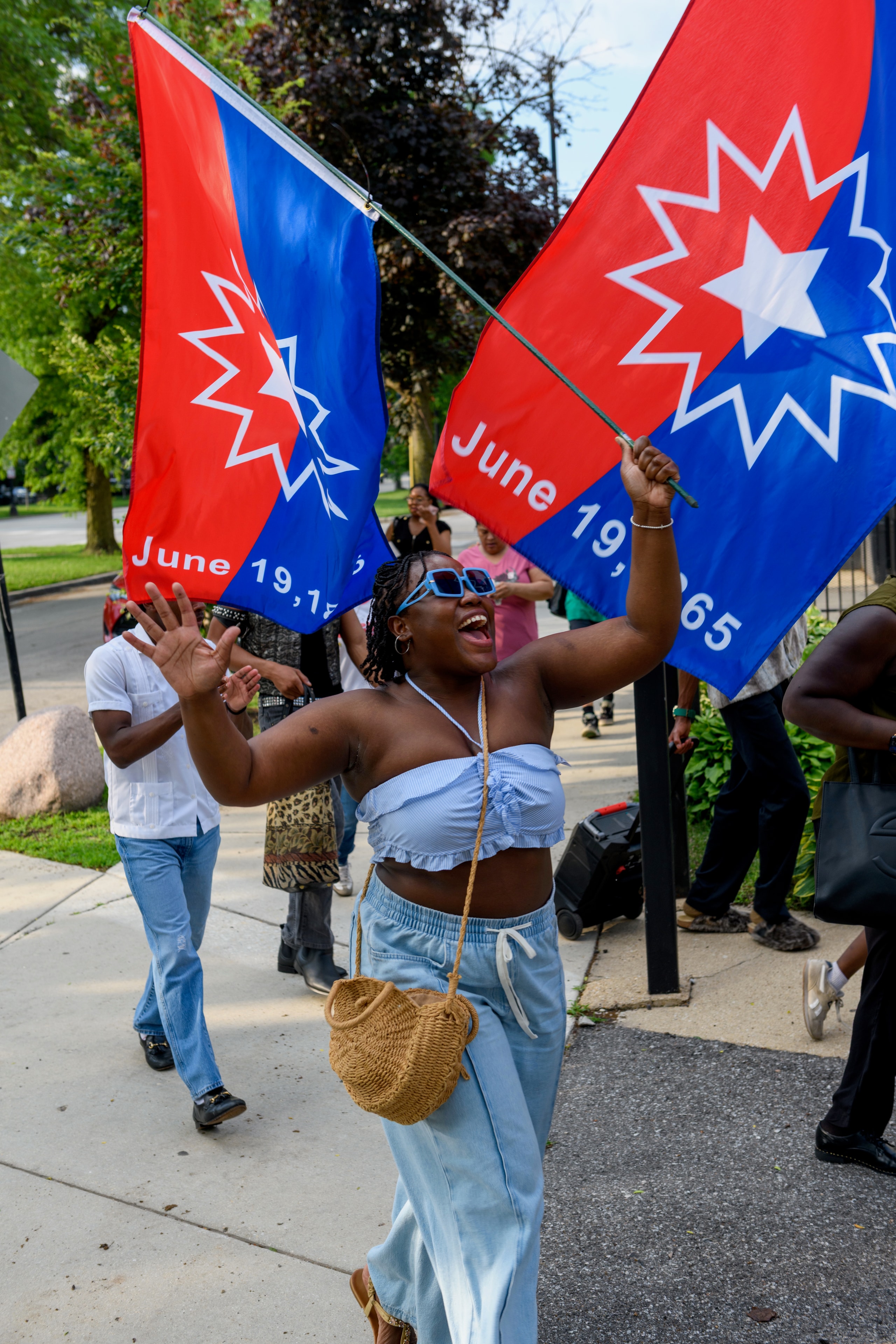 A Juneteenth parade participant waving red and blue Juneteenth flags marked June 19, 1865 along a sidewalk.